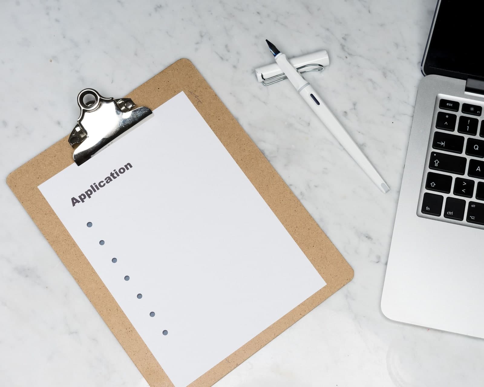 An application form on a clipboard beside a laptop and two pens on a marble desk
